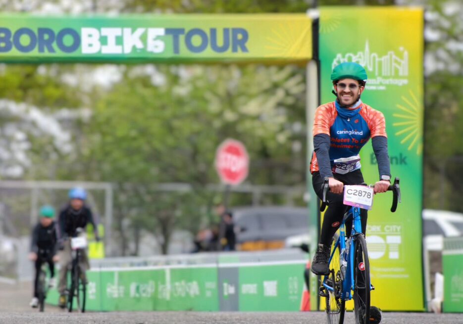 Cyclist wearing a CaringKind Athletes to End Alzheimer’s jersey rides in the TD 5 Boro Bike Tour, crossing the finish line at an outdoor event with other cyclists and event signage in the background.